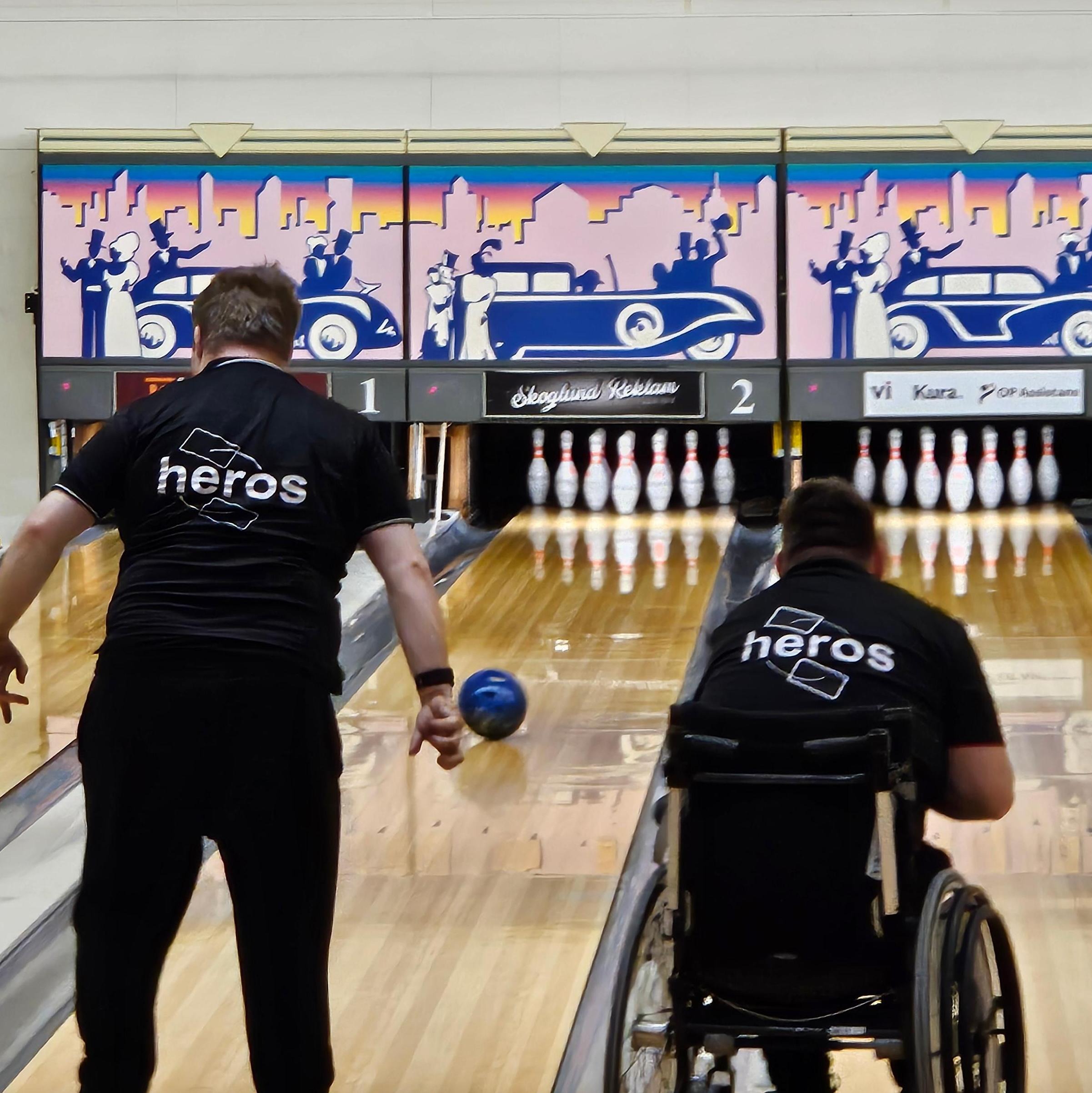 På bilden syns två personer som bowlar i en bowlinghall. Den ena personen står upp och är precis i rörelsen att släppa en blå bowlingklot mot banan. Den andra personen sitter i rullstol och bowlar på banan bredvid.