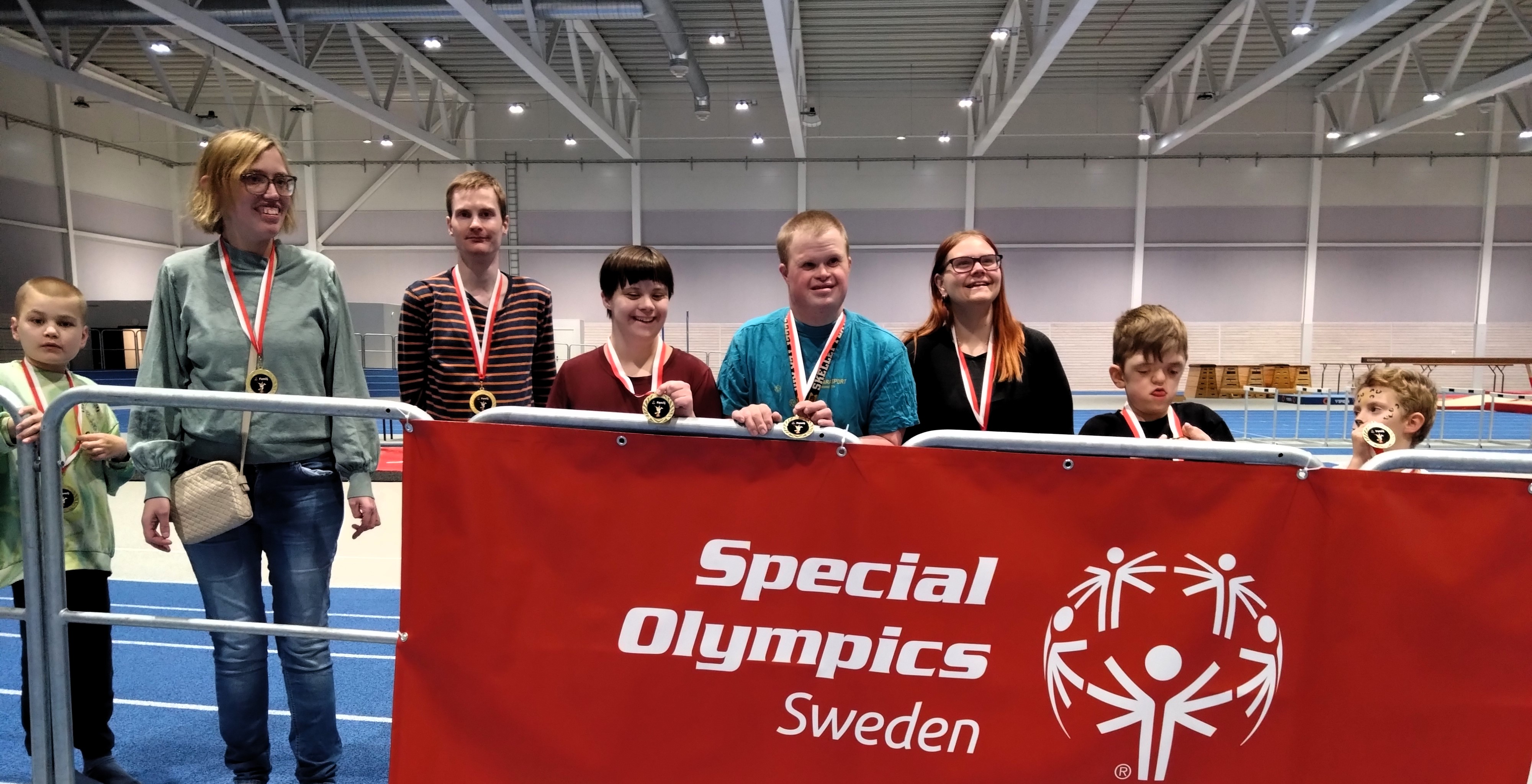 Åtta deltagare i varierande åldrar poserar med sina medaljer bakom Special olympics-flaggan