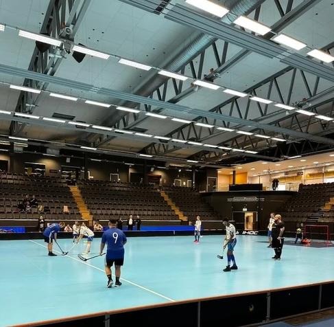 En innebandymatch i en stor inomhusarena där spelare står uppställda på den blå planen redo för spel och en läktare i bakgrunden.