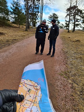 I förgrunden en uppförstorad orienteringskarta. I bakgrunden två personer som diskuterar vägval/orientering.