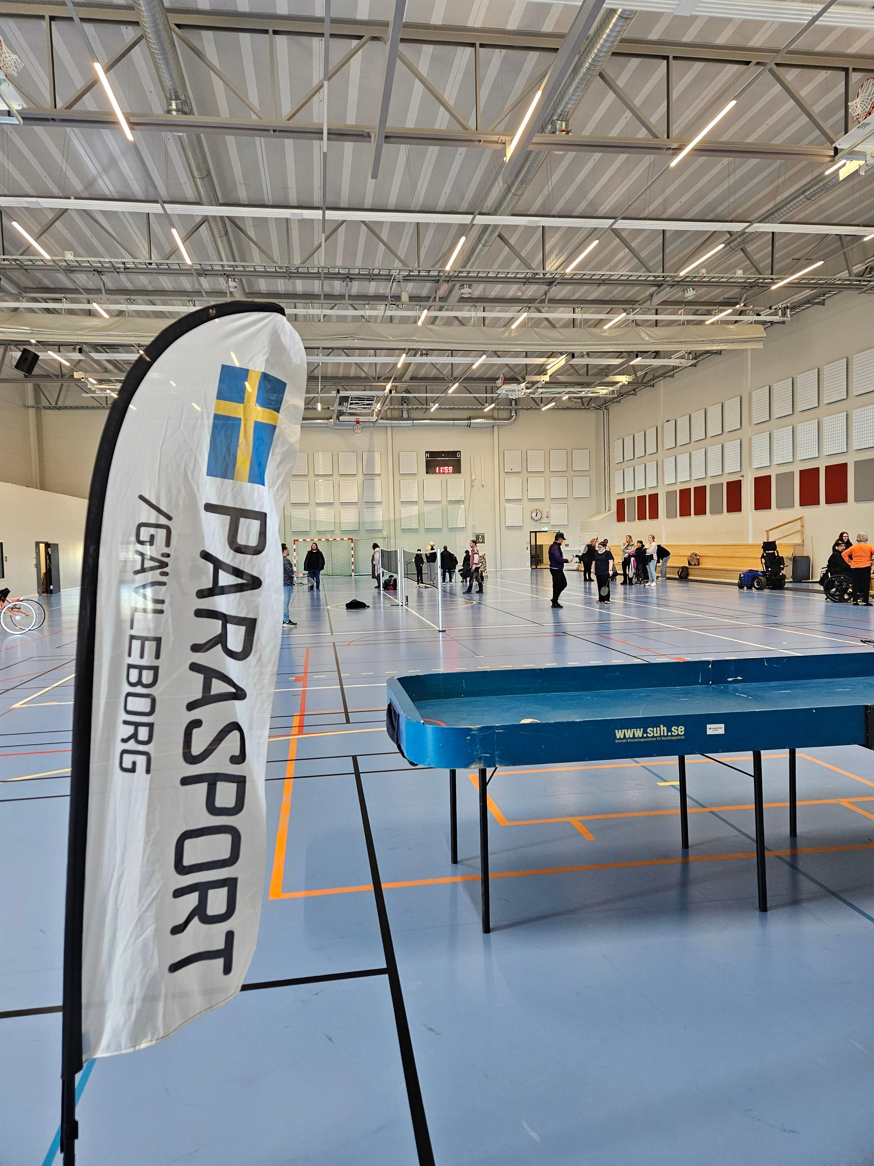 Beachflagga med Parasport Gävleborg och ett showdownbord i en idrottshall. Personer syns i bakgrunden.