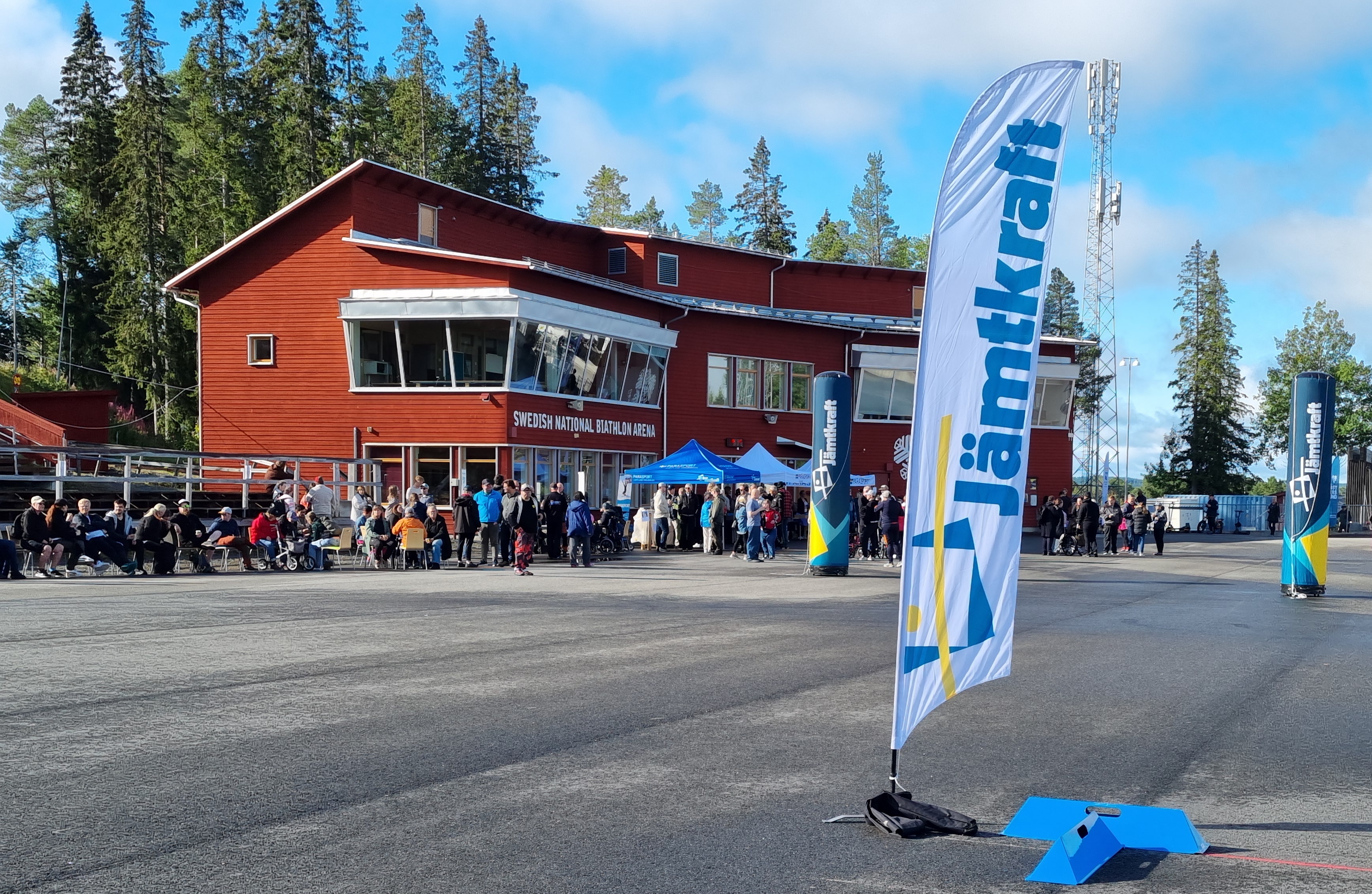 DV-dagen_I förgrunden en Jämtkraft beach-flag. Klarblå himmel. Mycket personer framför en röd byggnad "Swedish National Biathlon arena", när det är lunch-tid i det vackra vädret.