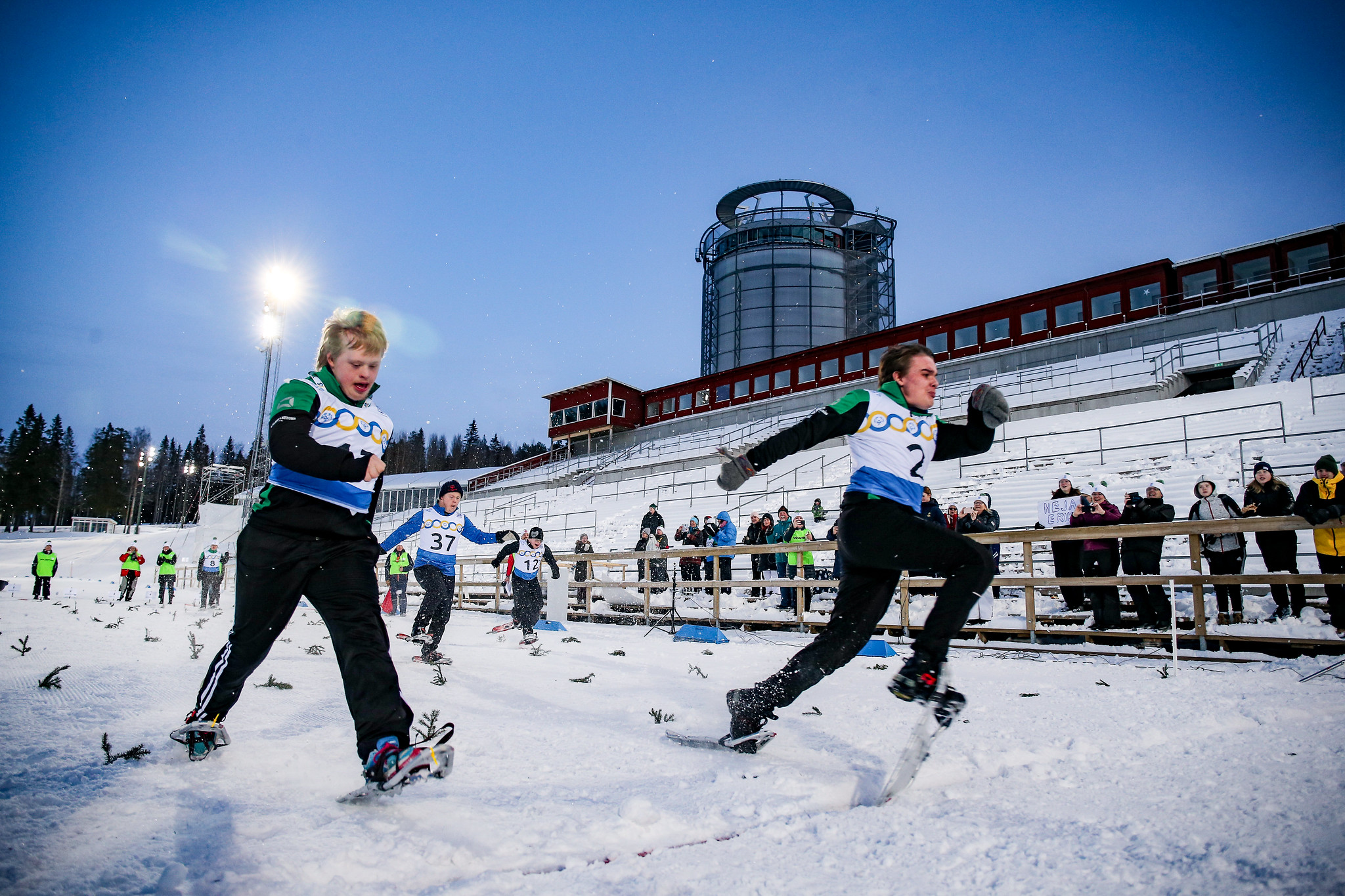 Utomhusmiljö, skidstadion. Två snöskolöpare är med stora steg på väg mot mål. I bakgrunden hejar publik. Det är kväll och strålkastarna lyser upp snön.