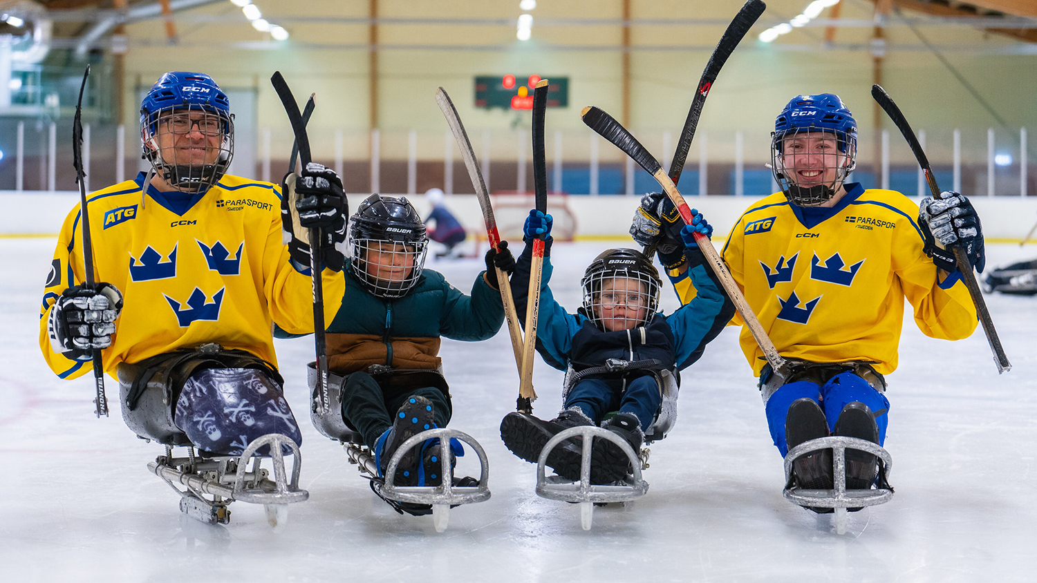 Fyra spelare uppradade på isen. Två unga nybörjare i mitten och två landslagsspelare på kanterna. Spelarna ler och sträcker armarna upp i luften.
