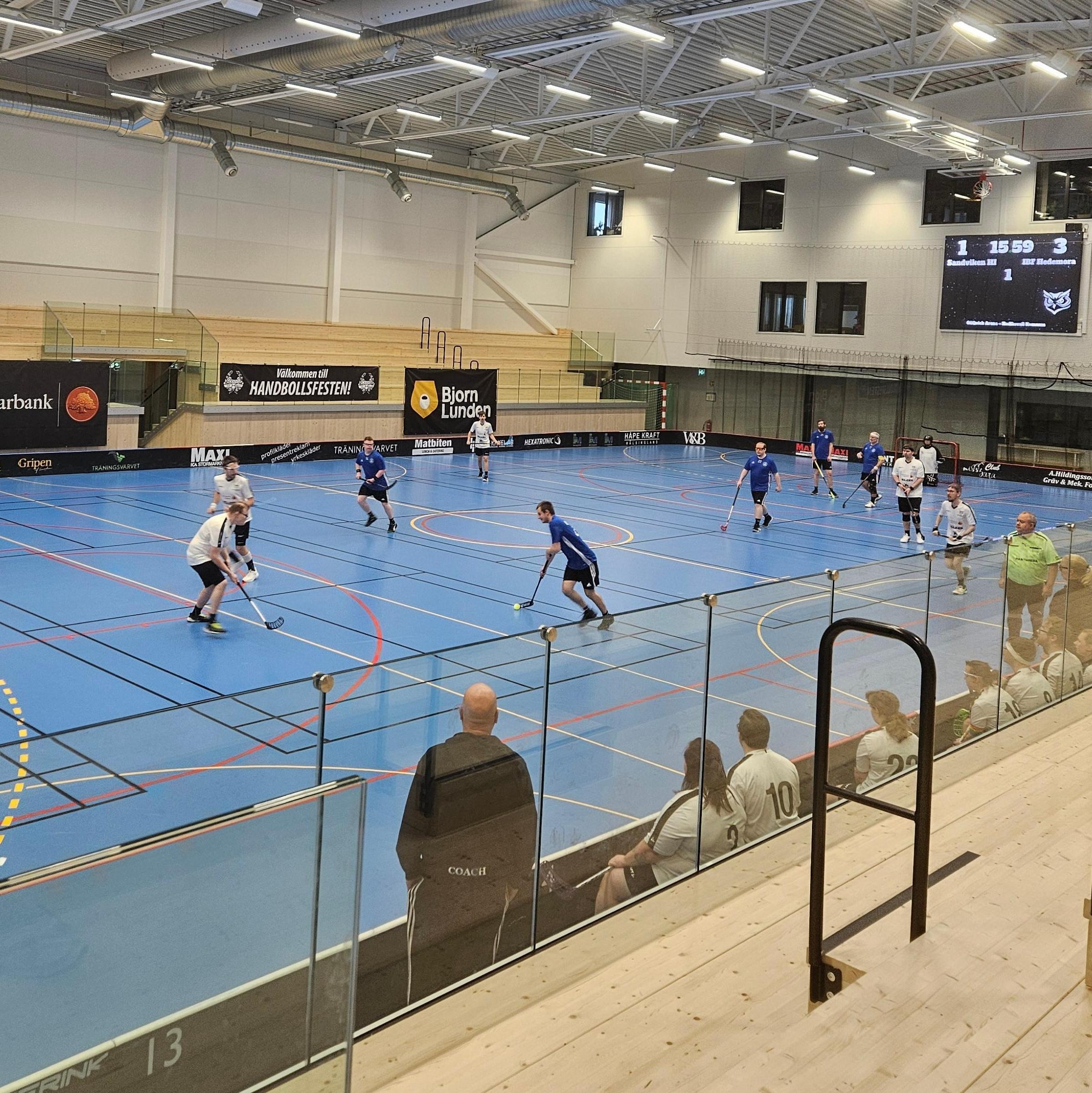 En innebandymatch pågår i en modern idrottshall med blått golv och högt i tak. Två lag, ett i blå tröjor och ett i vita, spelar på planen medan avbytare och ledare sitter på bänken i förgrunden. Några personer står vid sargen och följer spelet. På väggen syns en matchklocka och sponsorbanderoller.