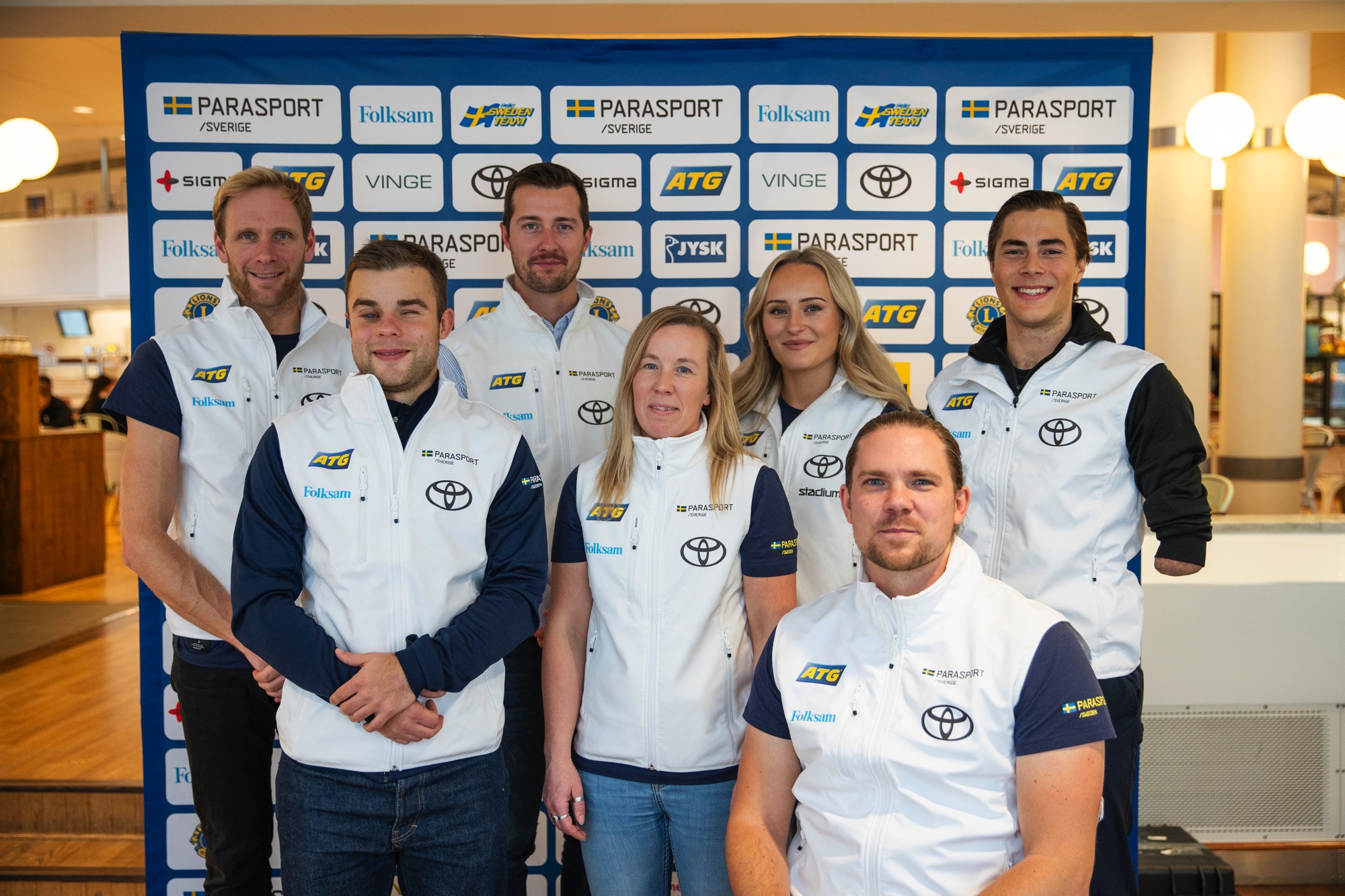 Gruppbild på paralandslagen i alpint och längd/skidskytte framför en sponsorvägg. Från vänster syns Daniel Richardsson, förbundskapten längd/skidskytte, Zebastian Modin, längd/skidskytte, Henrik Bergqvist, förbundskapten alpint, Nina Jonsson, förbundskapten längd/skidskytte, Ebba Årsjö, alpint, Arnt-Christian Furuberg, längd, Aaron Lindström, alpint.