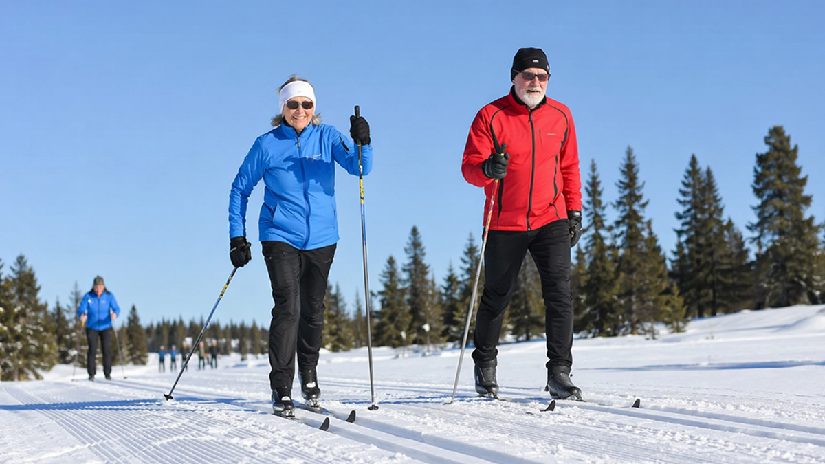 En äldre man och en äldre kvinna åker längdskidor på den kritvita snön. Himlen är klarblå.