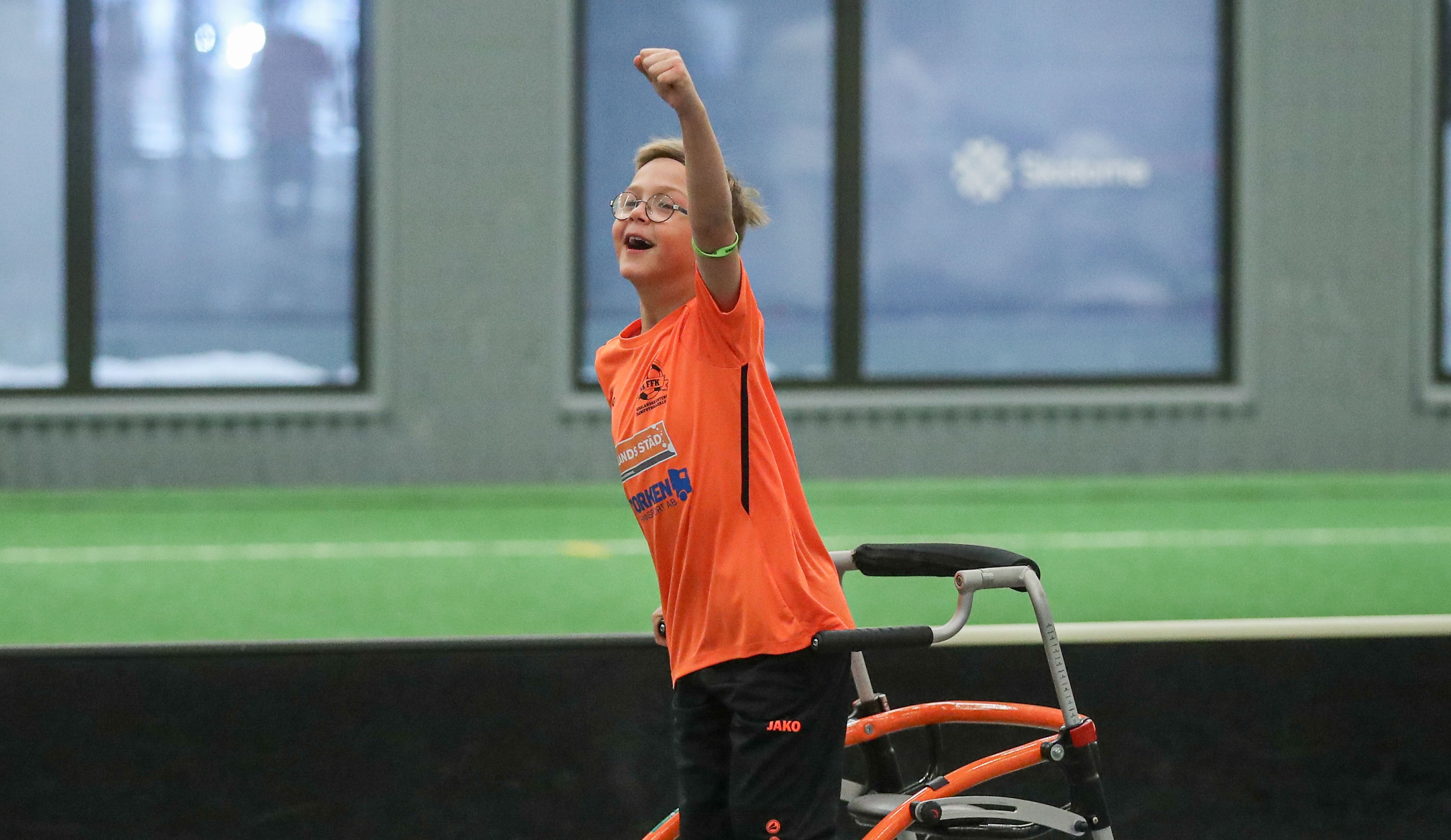 Foto, idrottshall. En pojke med gåstol håller leende upp en arm i luften som en segergest. Han har fotbollskläder på sig.