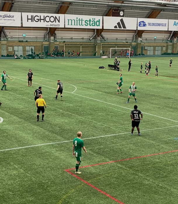 Bilden visar en inomhushall med konstgräs där en fotbollsmatch pågår. Det är ett 7–7-spel (eller liknande) och spelarna är utspridda över planen. Ett lag har gröna tröjor och det andra laget har mörka tröjor (svarta). I förgrunden syns en domare i gul tröja. I bakgrunden syns reklamskyltar längs med hallens väggar,