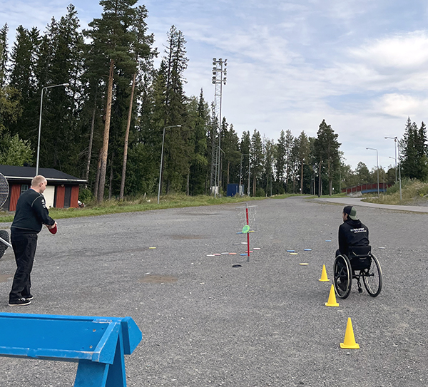 DV-dagen. Frisbeegolf. En kille står till vänster i bild och kastar frisbee mot trådkorgen. Till höger i rullstol sitter en instruktör.