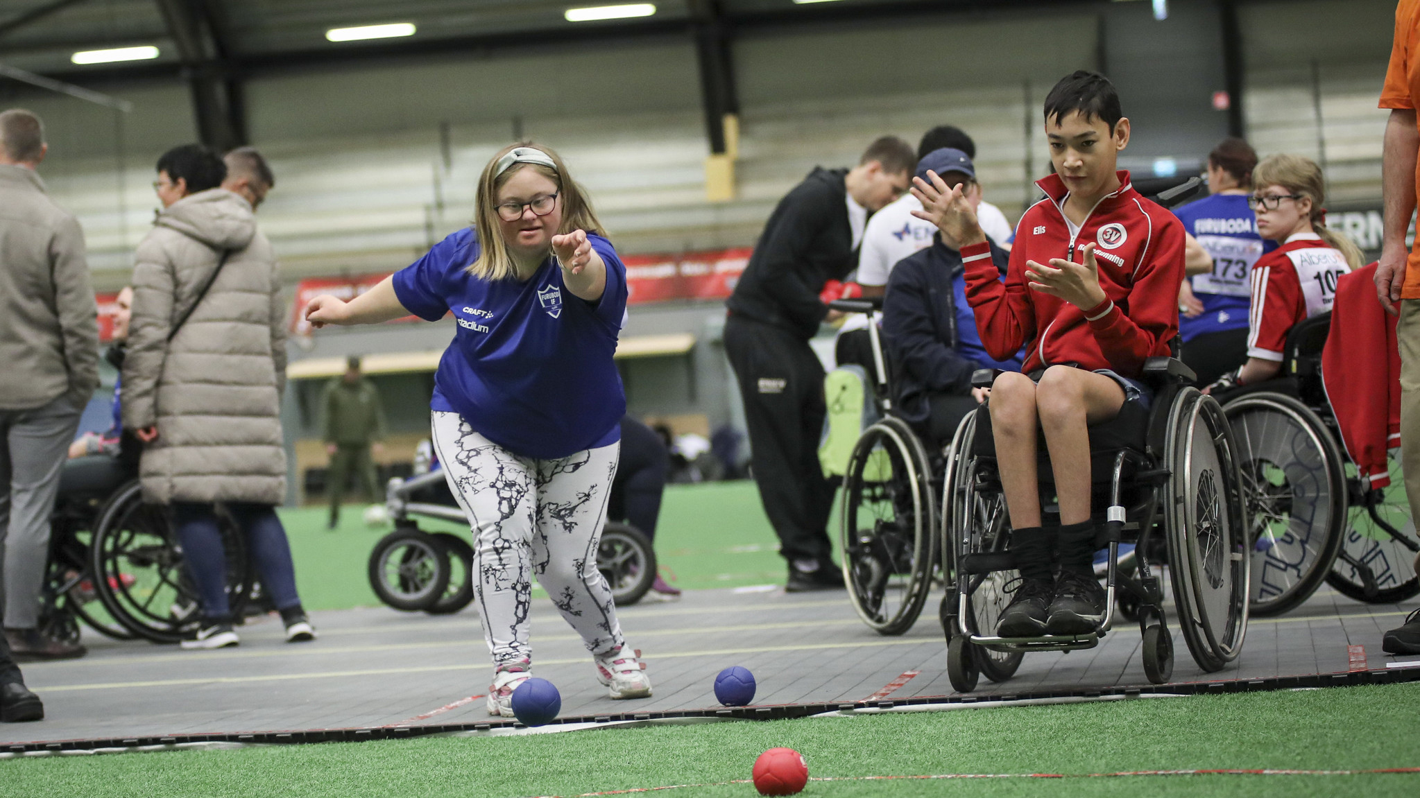Två personer spelar boccia. En tjej i blå tröja och en kille i röd tröja som spelar i rullstol.