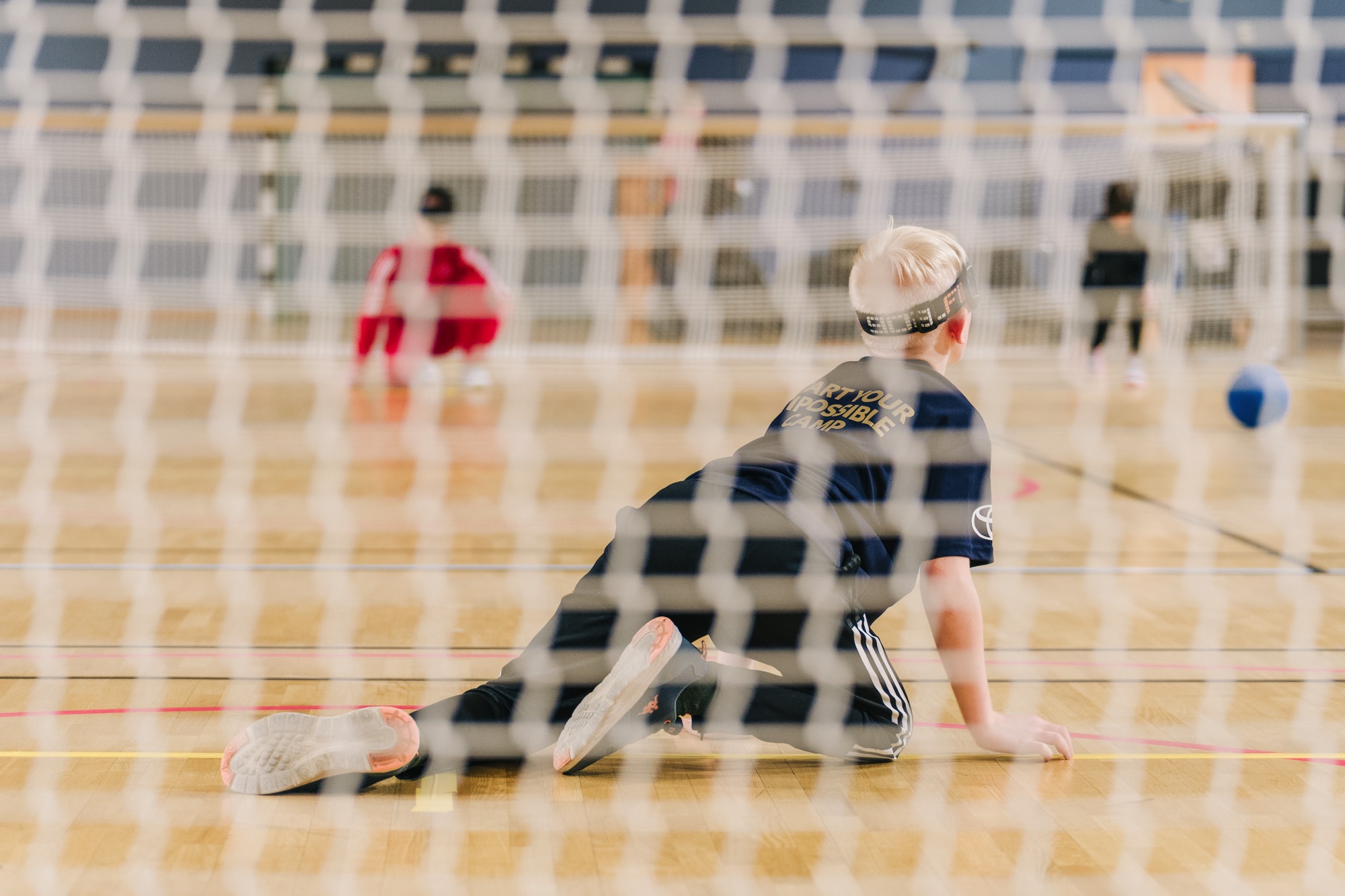Inomhus, idrottshall. Målnät i förgrunden, framför nätet en ung pojke som spelar goalball, sittande på golvet.