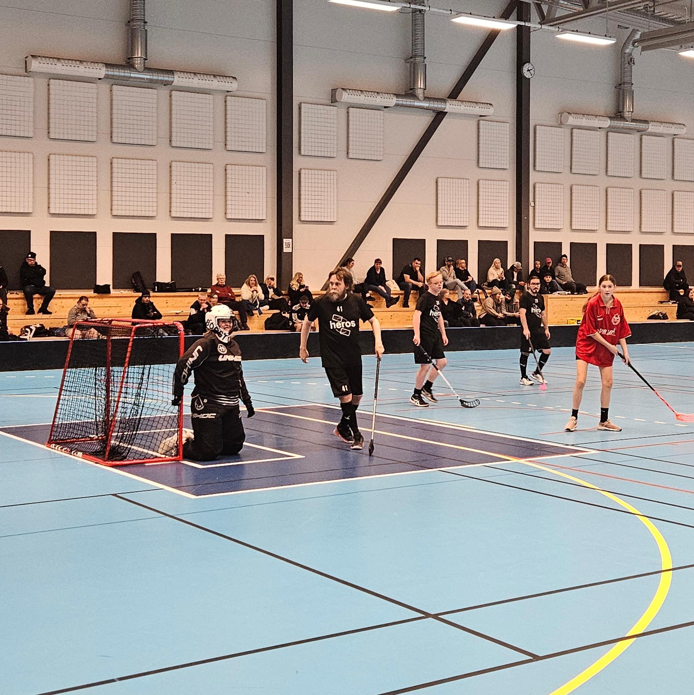Innebandymatch i en idrottshall där spelare är samlade runt målet på den blå planen, med publik och ledare i bakgrunden.