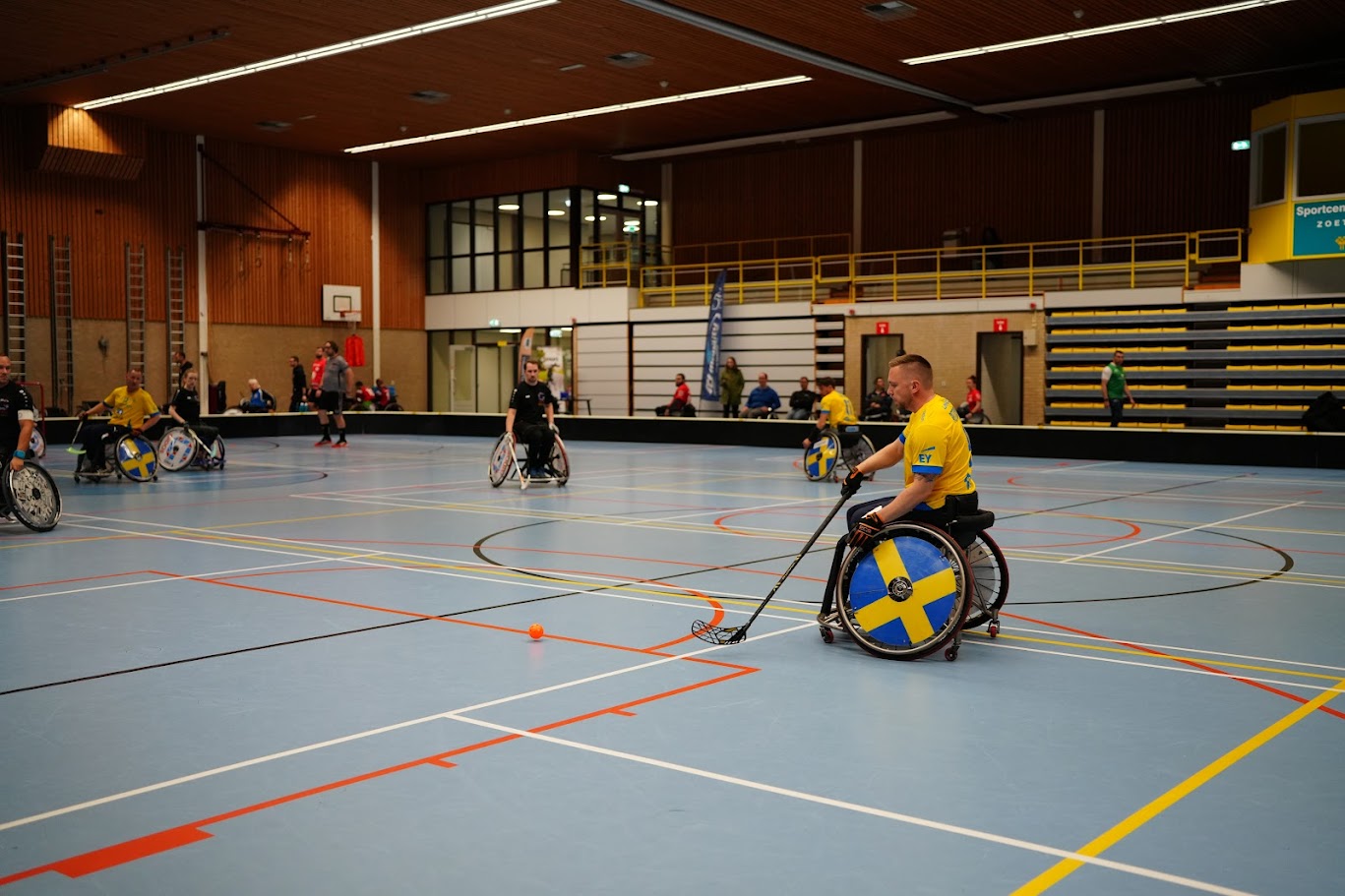En rullstolsinnebandyspelare Johnny Pettersson i en gul tröja och svensk flagga på rullstolens hjul är i fokus, på väg att slå till en orange boll med sin klubba. Flera andra spelare i sportrullstolar syns på det ljusblå golvet i sporthallen.