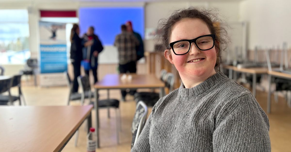 En glad tjej med mörka glasögon och grå tröja. I bakgrunden syns ett konferensrum med bord, stolar och några suddiga individer som står och pratar.
