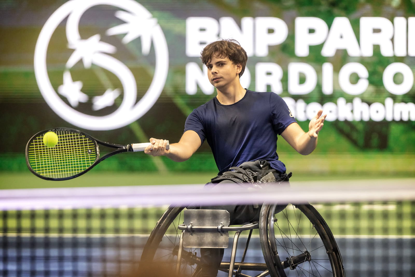 Foto, tennishall. Oscar i en rullstol, i färd med att svinga tennisracket mot bollen.