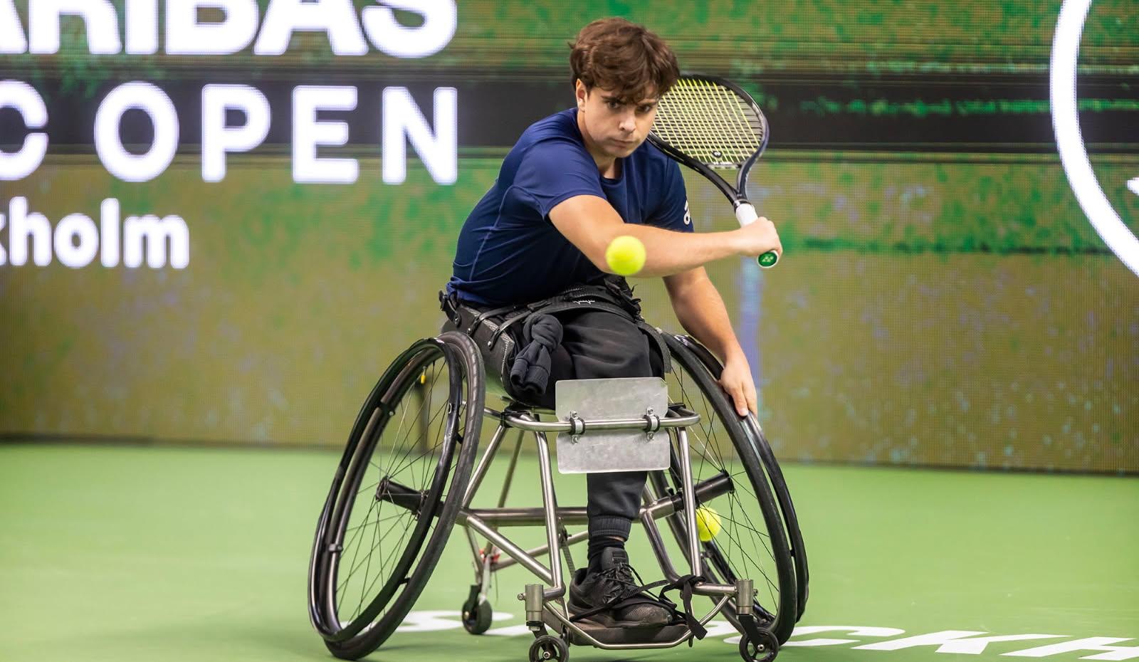 Foto, tennishall. Oscar i en rullstol, i färd med att svinga tennisracket mot bollen.